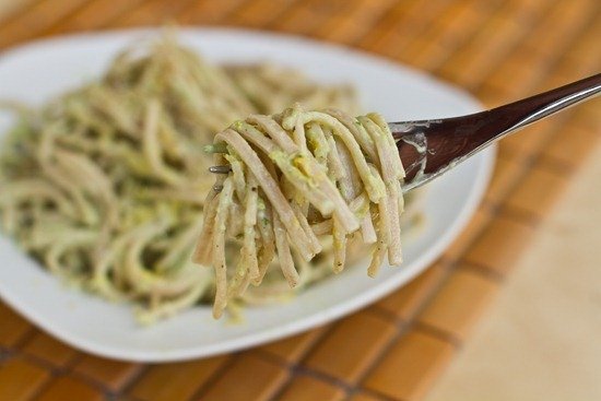 A fork holding a bite of creamy green pesto pasta, with more pasta on a white plate in the background, placed on a wooden surface.