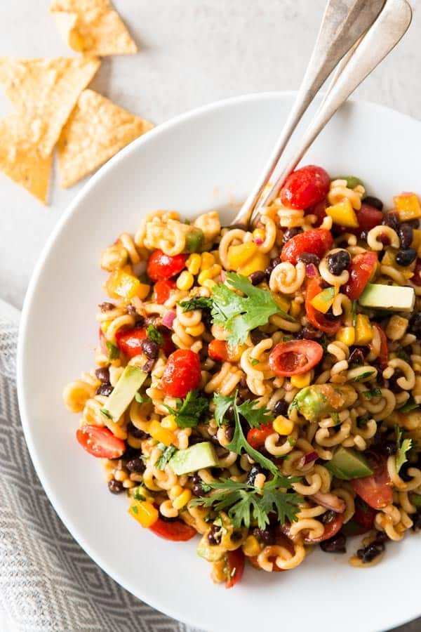 A white plate of pasta salad with cherry tomatoes, black beans, corn, diced peppers, and avocado, garnished with cilantro, with tortilla chips in the background.