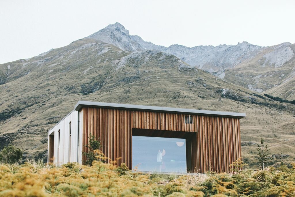 Modern house with wooden exterior, large front window, set in a mountainous landscape with dry grass and sparse trees.