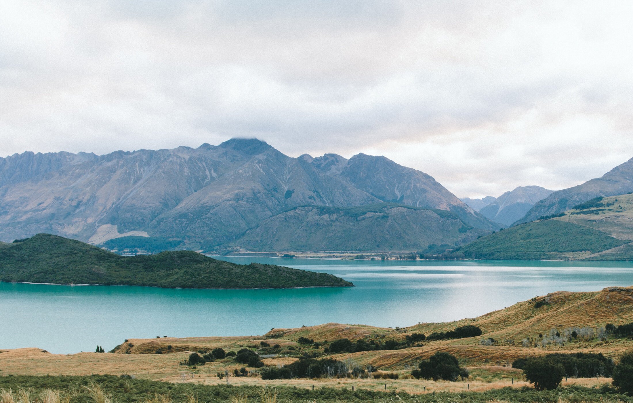 Scenic landscape of a lake with mountains in the background, green hills in the foreground, and cloudy sky.