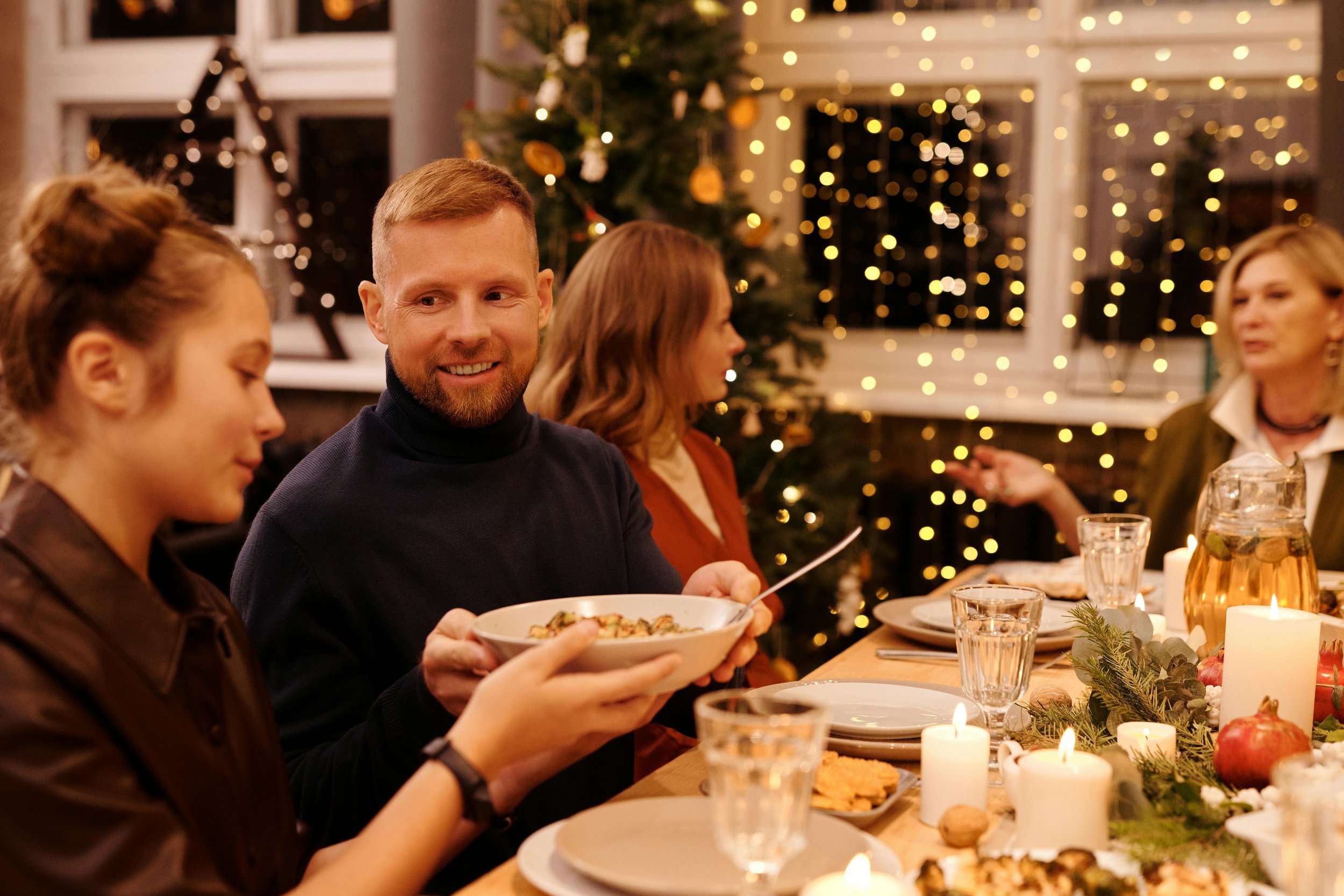 A group of four people enjoying a Christmas dinner at a decorated holiday table with candles, pomegranates, and greenery, in a warmly lit room with Christmas lights and a decorated tree in the background.