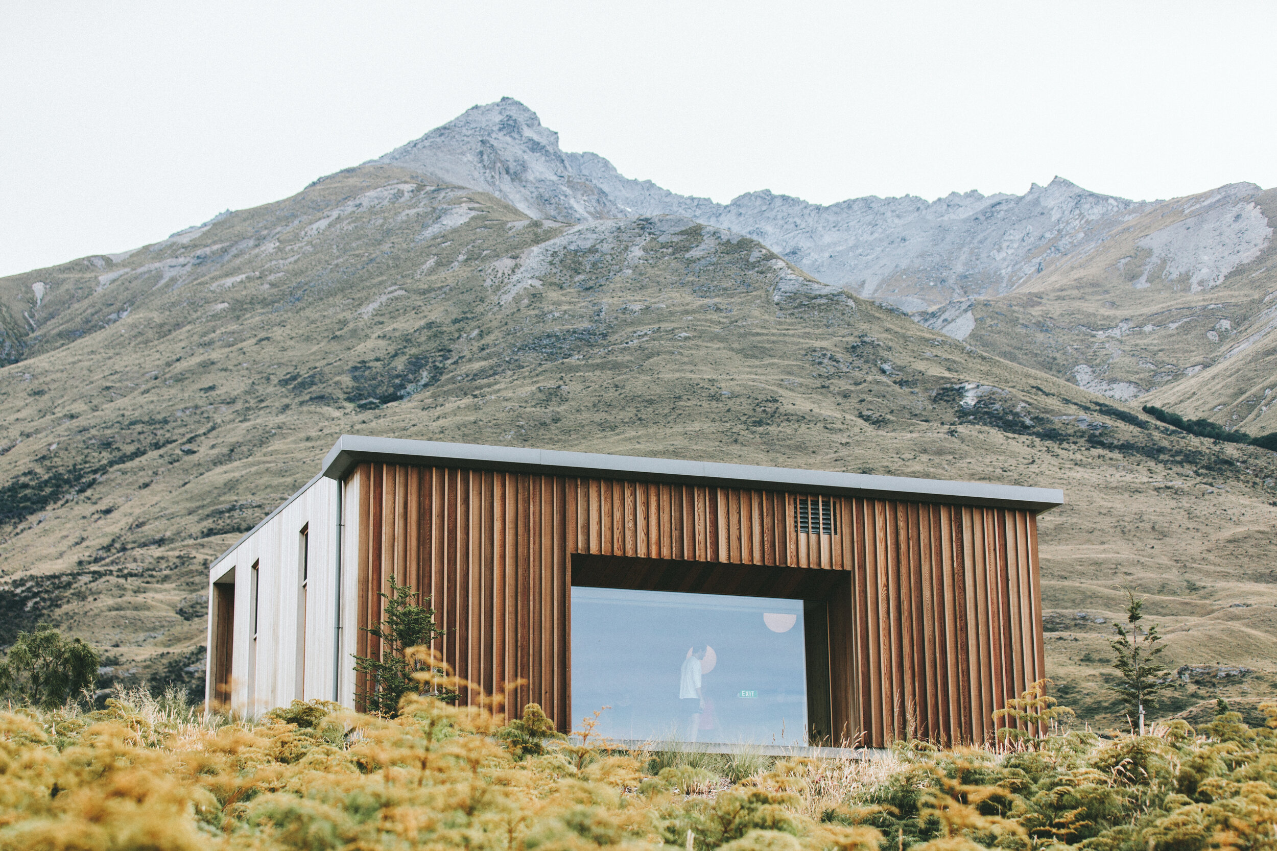 Modern house with wooden exterior and large glass window, set against a mountainous landscape with sparse vegetation and rocky peaks.