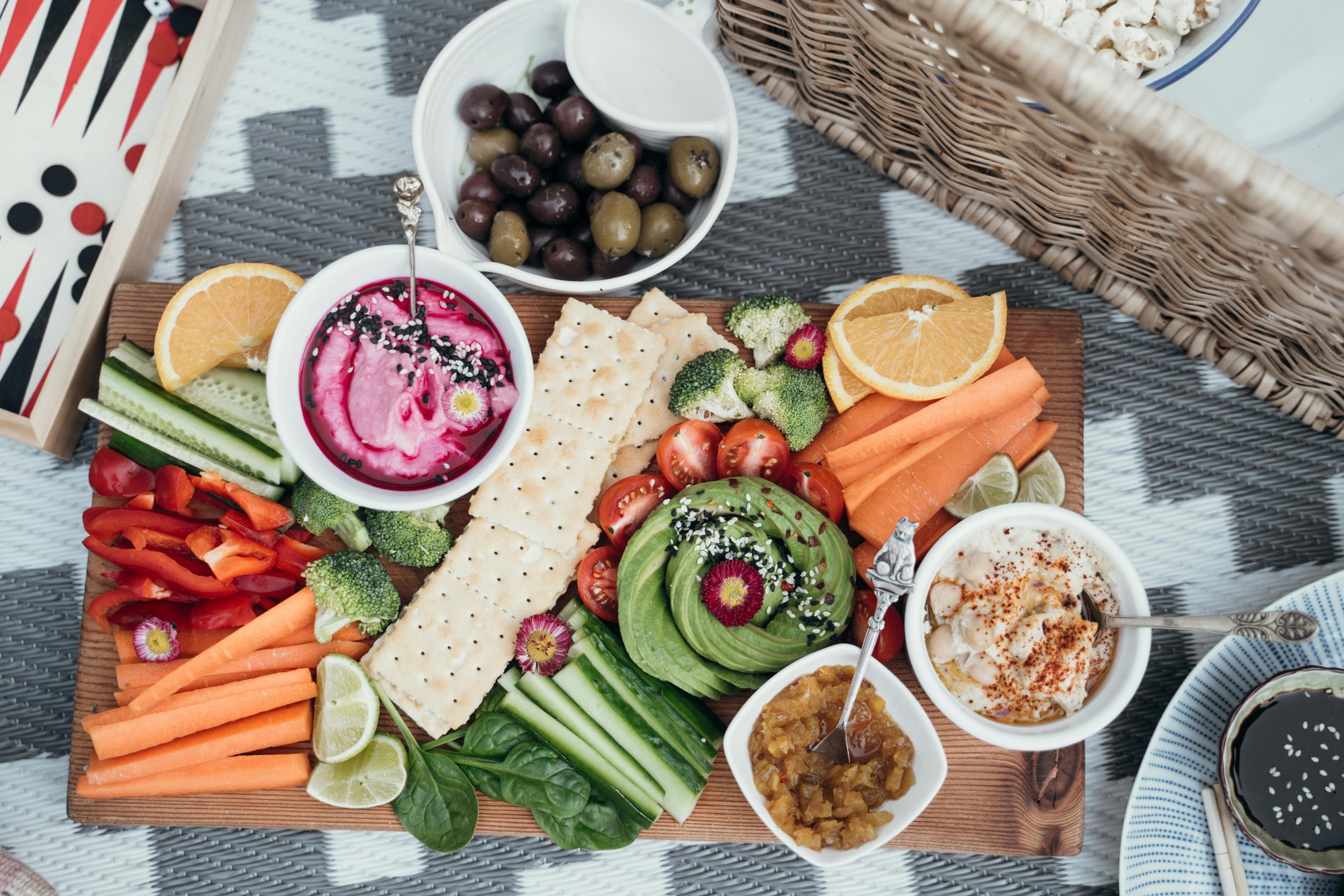 Assorted fresh vegetables, fruits, and dips arranged on a wooden platter, including cherry tomatoes, avocado, cucumber, carrots, broccoli, red bell peppers, lemon slices, crackers, olives, and bowls of colorful dips and spreads.