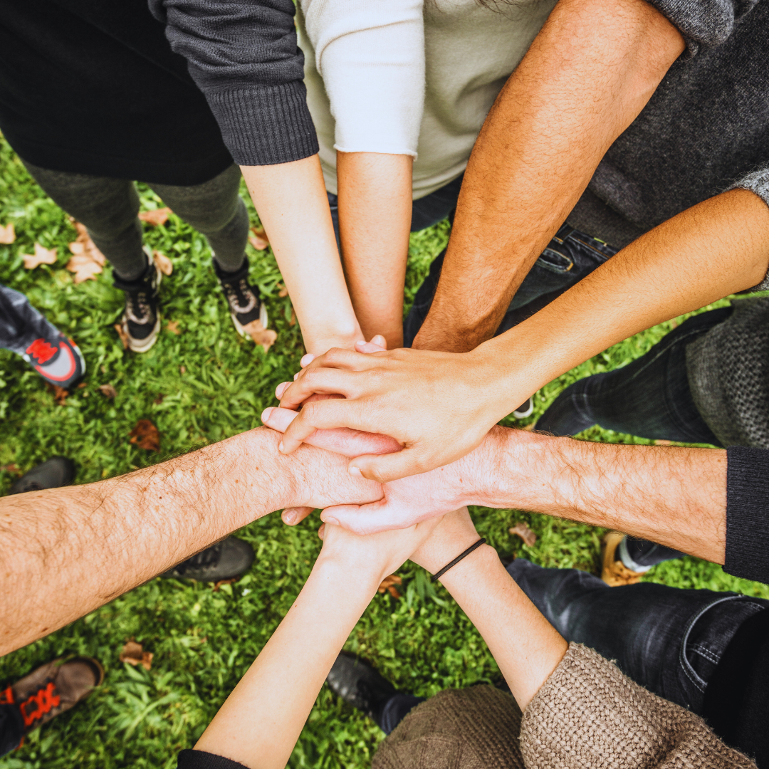 Multiple people placing their hands together in a circle, symbolizing teamwork or unity, on a grassy outdoor area.