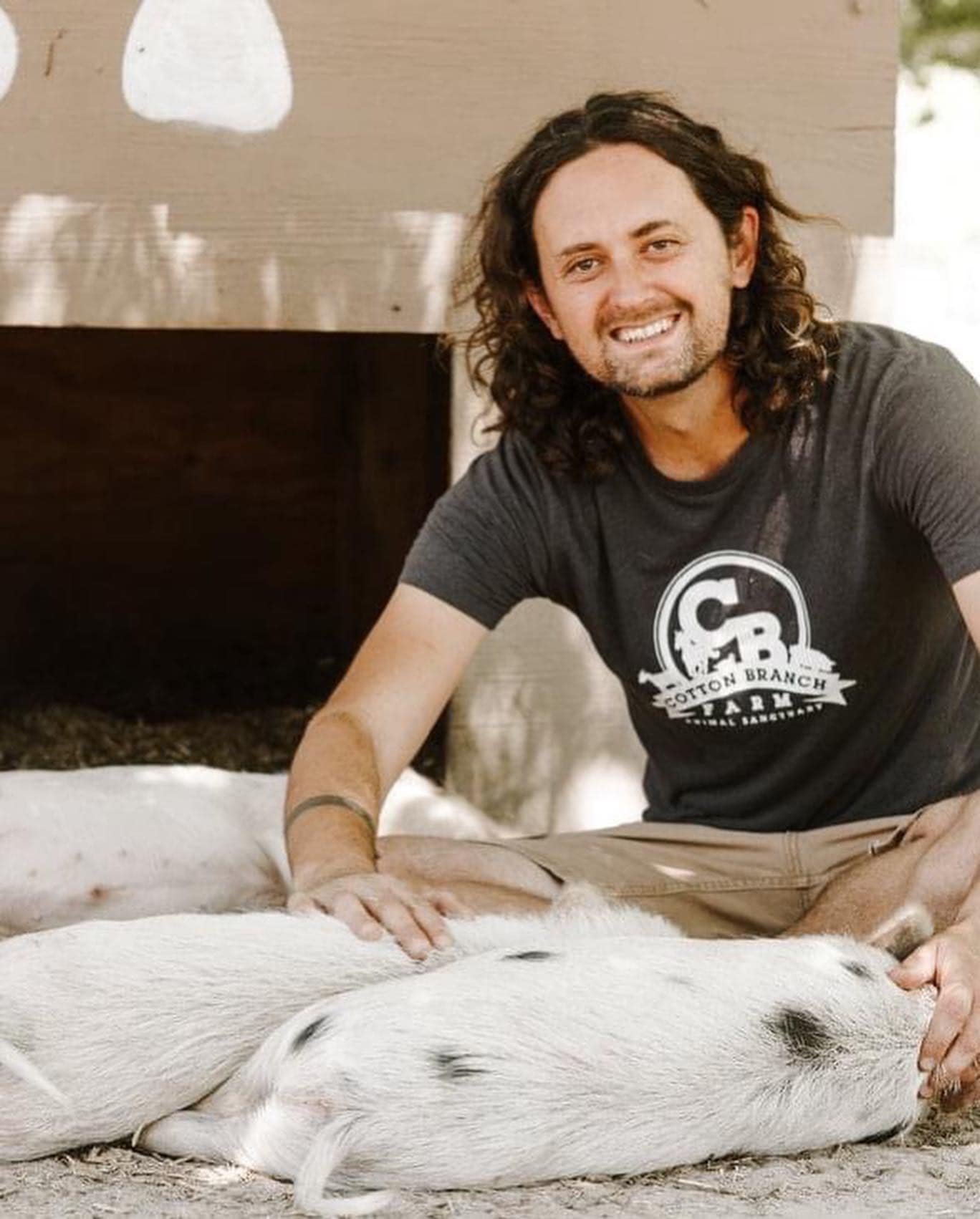 A man with long curly hair and a beard, smiling, sitting on the ground with a piglet lying on its side. The man is gently petting the piglet.