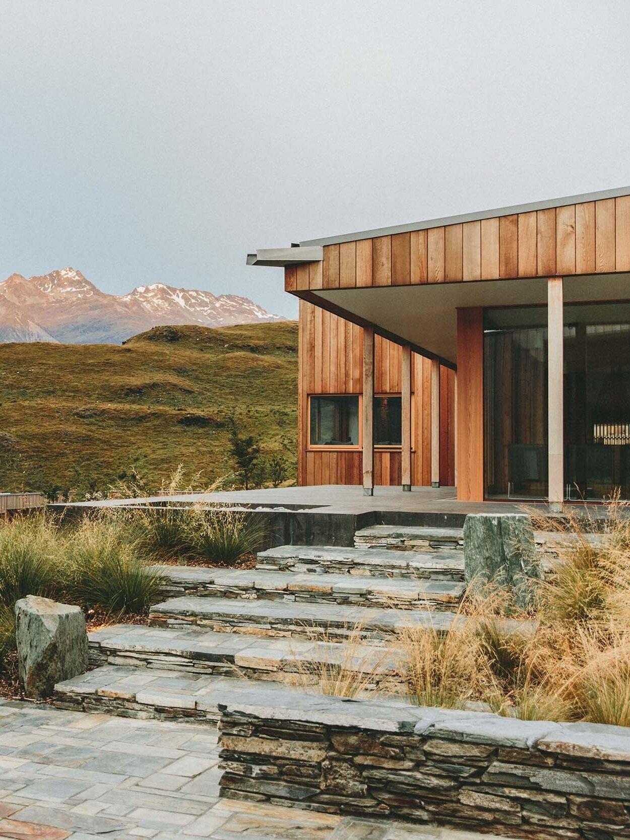 Modern house with wooden exterior and large glass windows, set in a mountainous landscape with snow-capped peaks and rolling hills, featuring stone steps and a stone patio in the foreground.