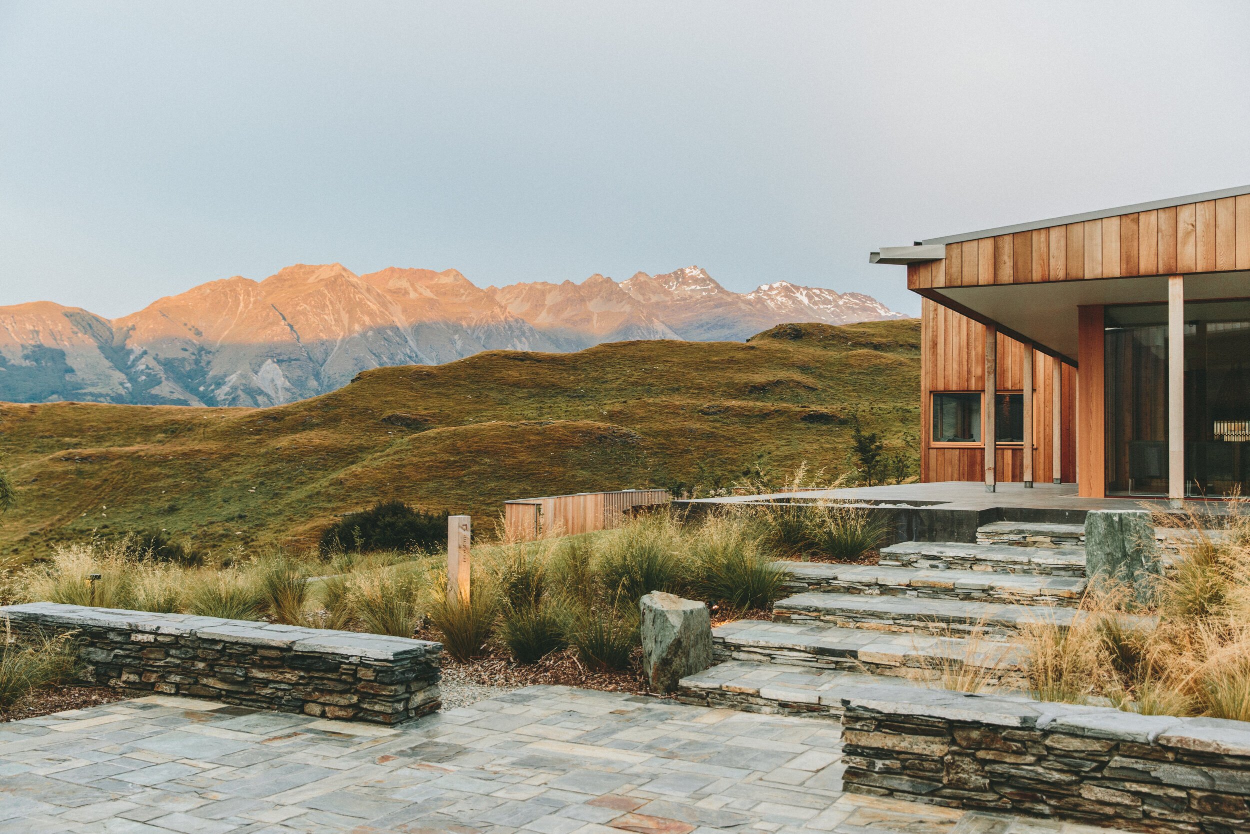 A modern house with wooden exterior in a landscape with rolling green hills and mountains in the background at sunset.