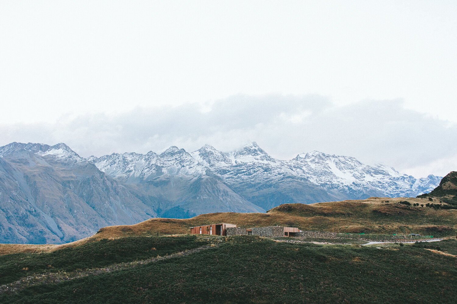 A mountain landscape with snow-capped peaks in the background and grassy hills in the foreground. There are modern buildings on the hillside.